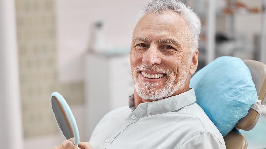 Smiling older man in the dental chair holding a hand mirror